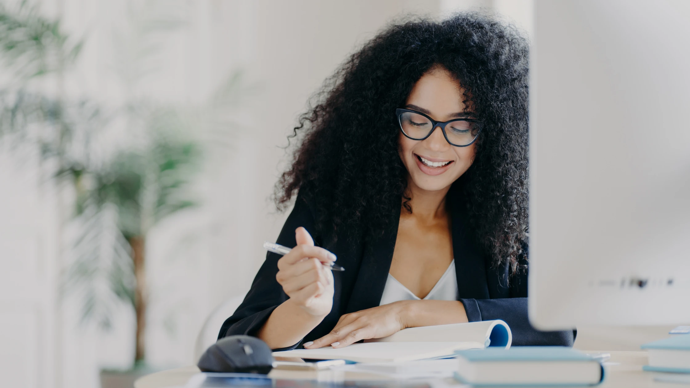A smiling woman with curly hair and glasses, reviewing study materials at her desk, representing a medical student preparing for the USMLE Step 1 exam using various resources like the AMBOSS Step 1 Self-Assessment.